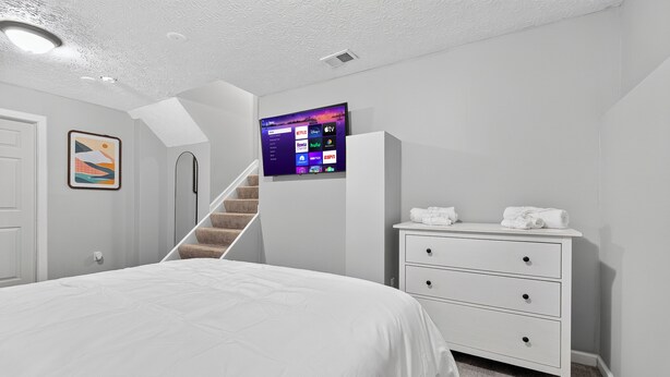 A bedroom with a bed, a dresser, a TV mounted on the wall, a staircase, and a door. - Elegant 3BR 5-Bed Spacious Home: Pool & Patio (Galloway)