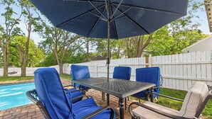An outdoor patio with a pool, surrounded by a white fence, featuring a table and chairs with blue cushions. - Elegant 3BR 5-Bed Spacious Home: Pool & Patio (Galloway)