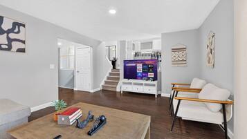 A spacious living area with a flat-screen TV, a coffee table with books and a remote, a white sofa, and a staircase leading to an upper level.