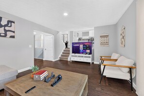 A spacious living area with a flat-screen TV, a coffee table with books and a remote, a white sofa, and a staircase leading to an upper level. - Elegant 3BR 5-Bed Spacious Home: Pool & Patio (Galloway)