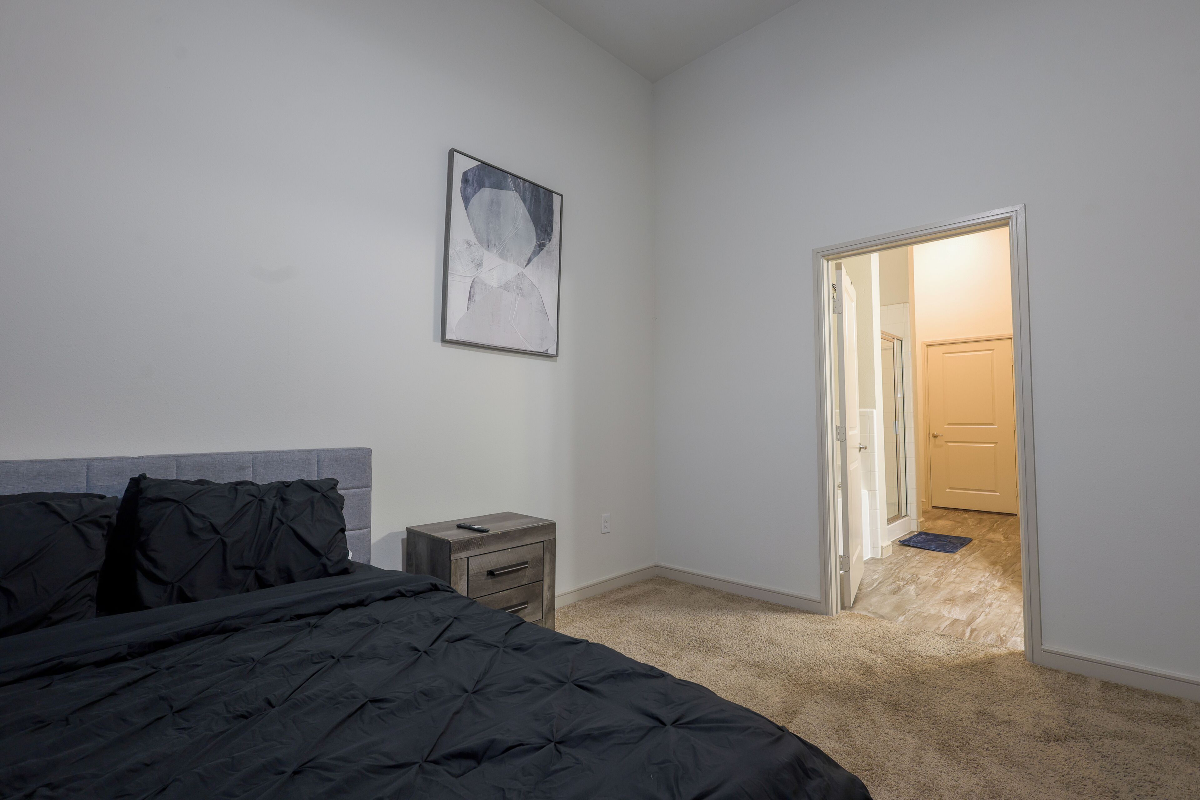 A bedroom with a bed, a nightstand, a wall-mounted picture, and an open door leading to another room.