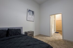 Een slaapkamer met een bed, nachtkastje, een aan de muur hangend schilderij en een open deur naar een andere kamer.