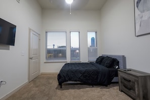 A bedroom with a bed, a dresser, a TV, and three windows with blinds.