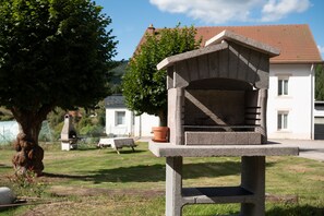 A stone birdhouse with a sloped roof, a concrete bench, and a potted plant in a grassy area with a tree and a house in the background.