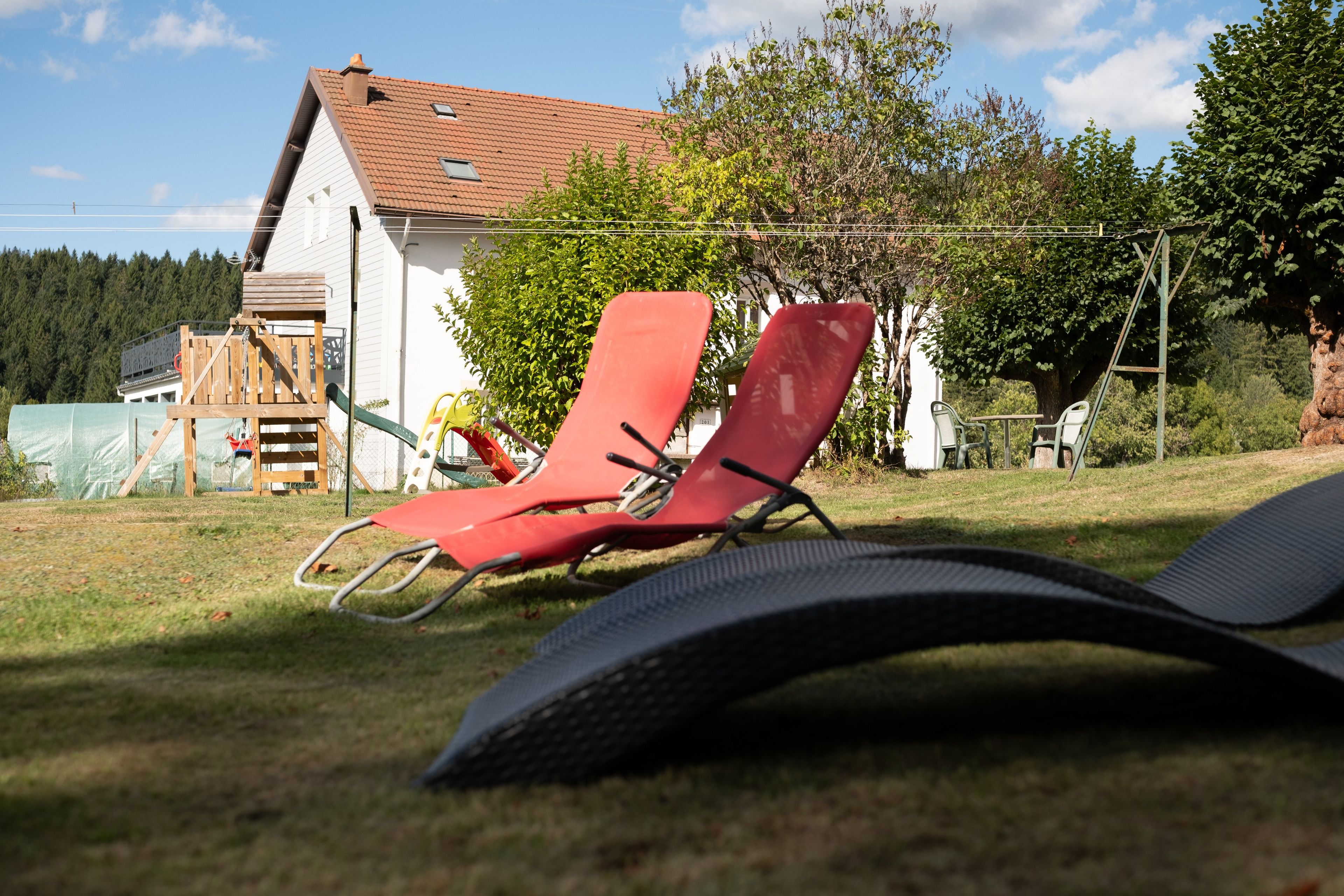 Een gazon met twee rode ligstoelen en een zwarte mat, een houten speelconstructie, en een wit gebouw met een rood dak op de achtergrond.