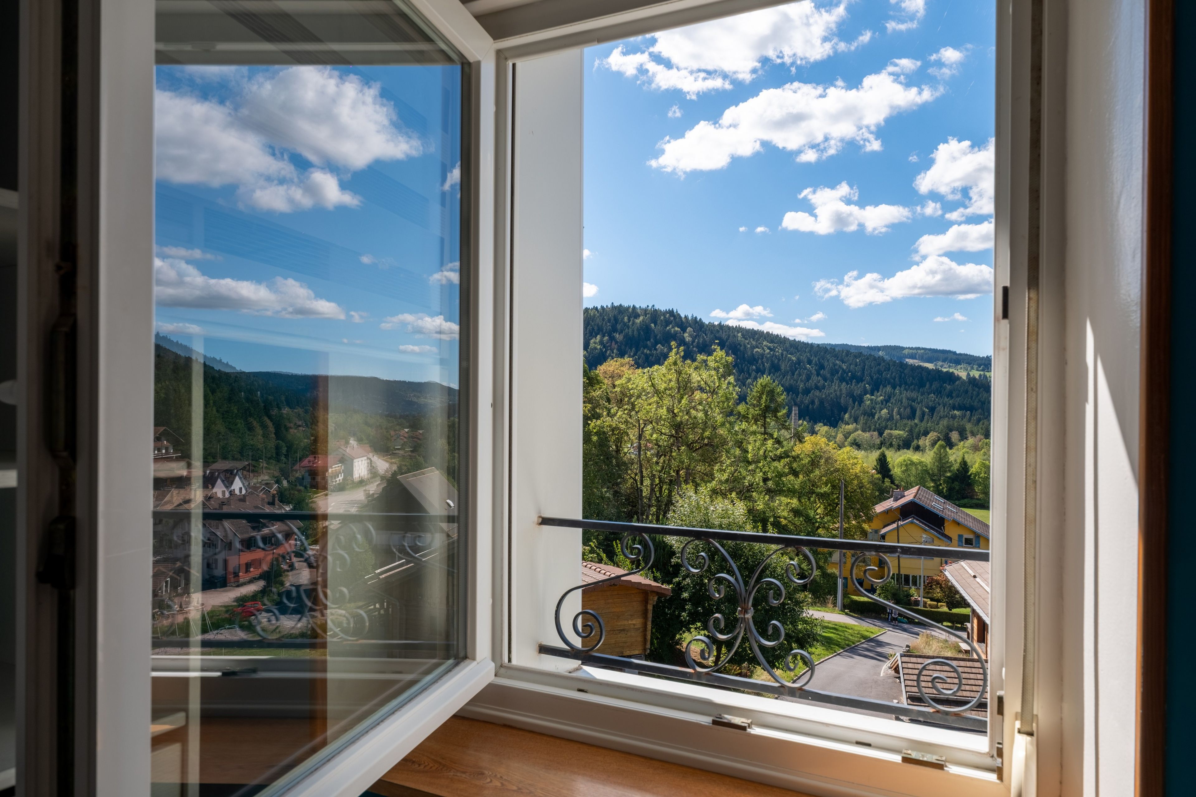 Une vue depuis une fenêtre ornée d’une balustrade en métal, donnant sur une vallée parsemée d’arbres et sur une chaîne de montagnes lointaine.