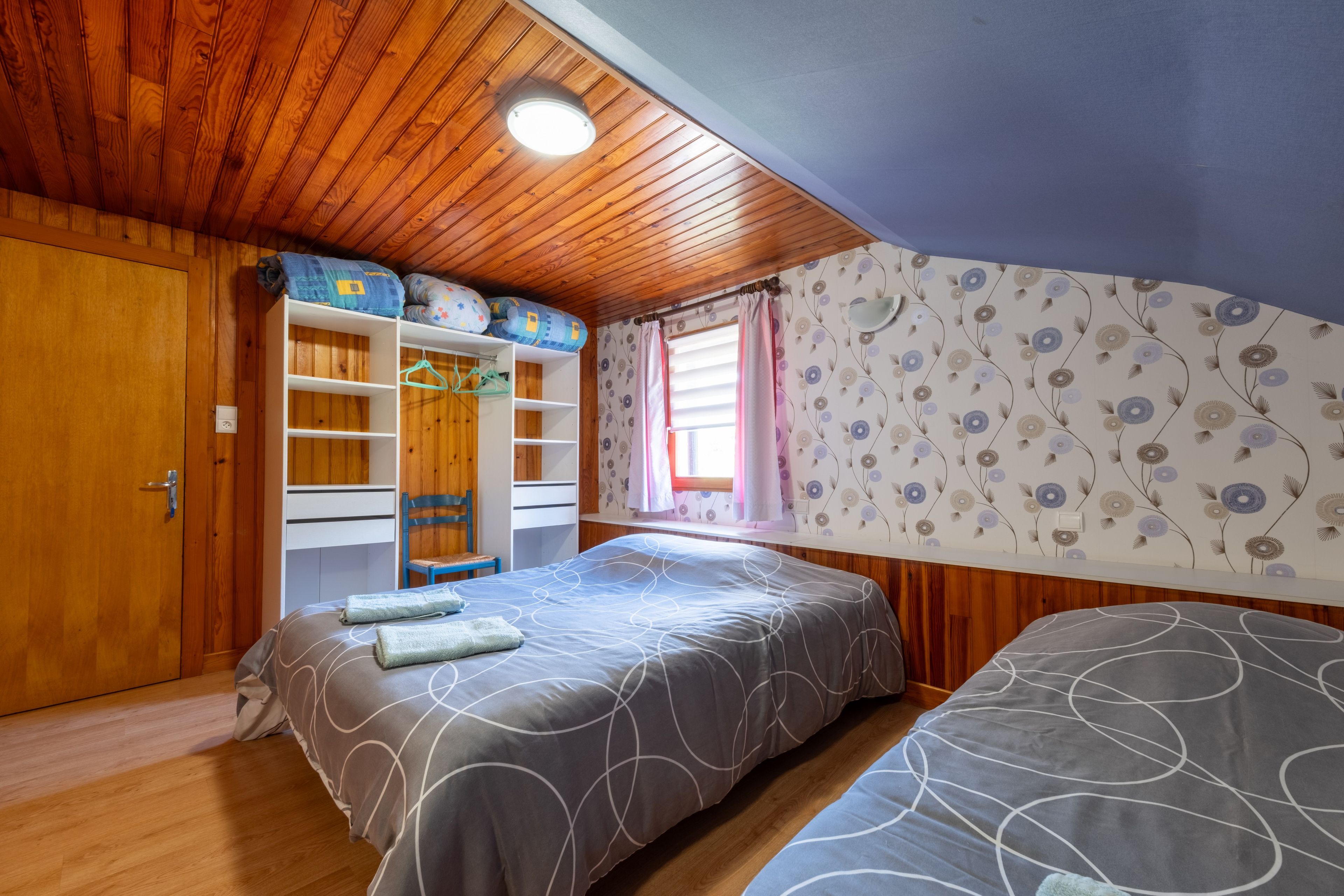 Ein Schlafzimmer mit Holzboden, einem Bett mit grau gemustertem Bettbezug, einem Kleiderschrank und einem Fenster mit floralem Wandpapier.