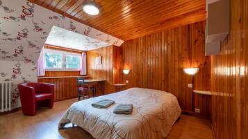 Ein Zimmer mit Holzwänden, einem Bett mit gemustertem Überwurf, einem roten Sessel, einem Tisch und einem Fenster mit Vorhängen.