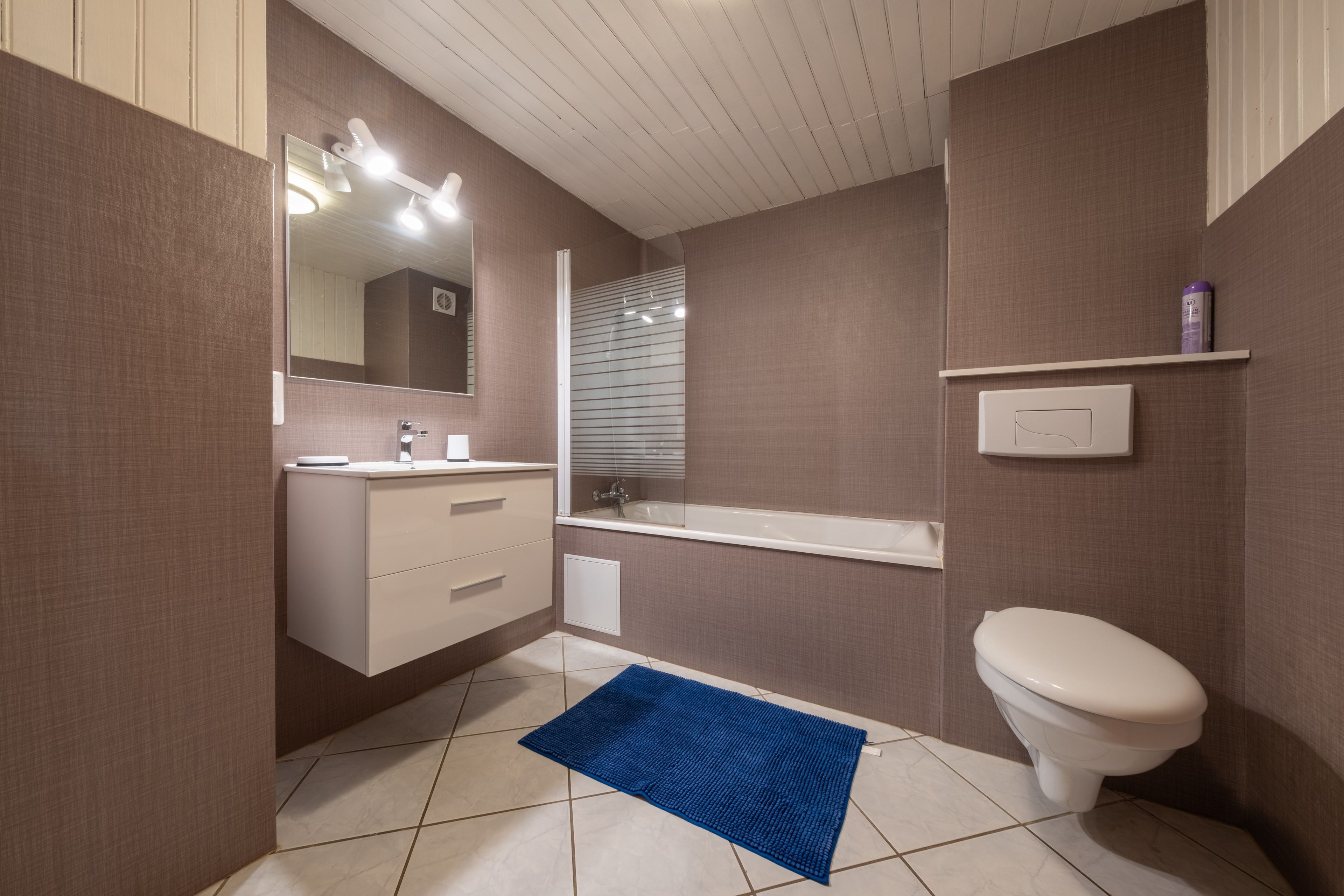Une salle de bain avec des toilettes blanches, un lavabo avec un miroir et un tapis de bain bleu.
