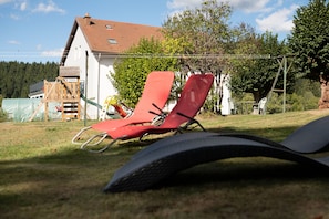 Des fauteuils de jardin et une structure de jeu en bois se trouvent dans une cour verdoyante, avec une maison en arrière-plan.