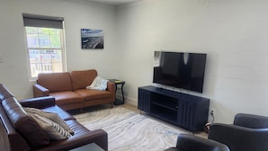 A living room with a brown sofa, a black armchair, a TV on a stand, a ceiling fan, and a window with a view of a building. - Charming 2-bedroom house in Oak Island close to the beach (Oak Island)