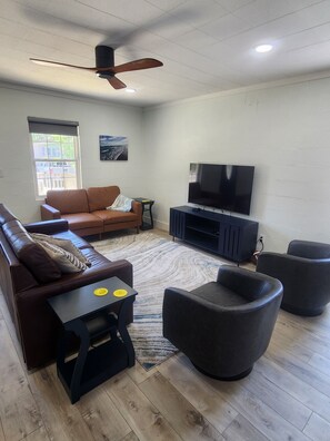 A living room with a brown sofa, a black armchair, a TV on a stand, a ceiling fan, and a window with a view of a building.