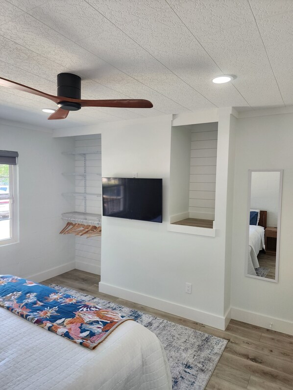 Un dormitorio con cama, ventilador de techo, televisor montado, espejo y ventana.