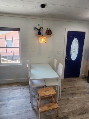 Una habitación con una mesa y sillas blancas, un taburete de madera, una lámpara colgante, una puerta azul con un panel de vidrio y una ventana que da a un edificio.
