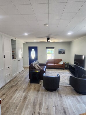 A spacious living room with a wooden floor, a ceiling fan, a television, and a large window. - Charming 2-bedroom house in Oak Island close to the beach (Oak Island)