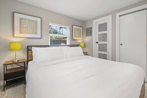 A bedroom with a large bed, two bedside tables with lamps, a window with blinds, a door, and framed artwork on the wall. - Perfect stay for 2-Walk to Wilton Drive-Shops &bars (Wilton Manors)