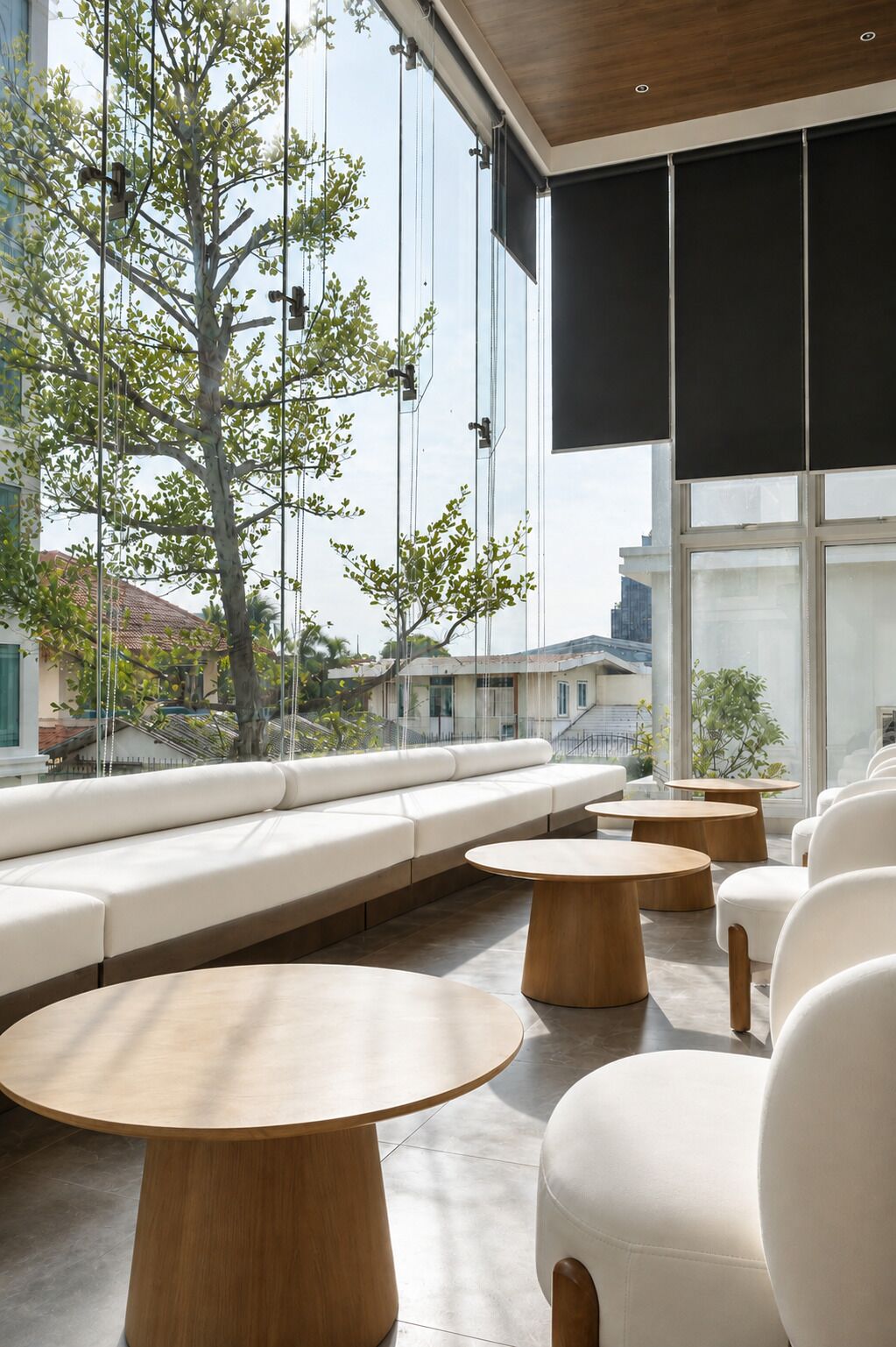 Zona relax moderna con un divano bianco, tavolini rotondi in legno e una grande finestra con vista su edifici e alberi.