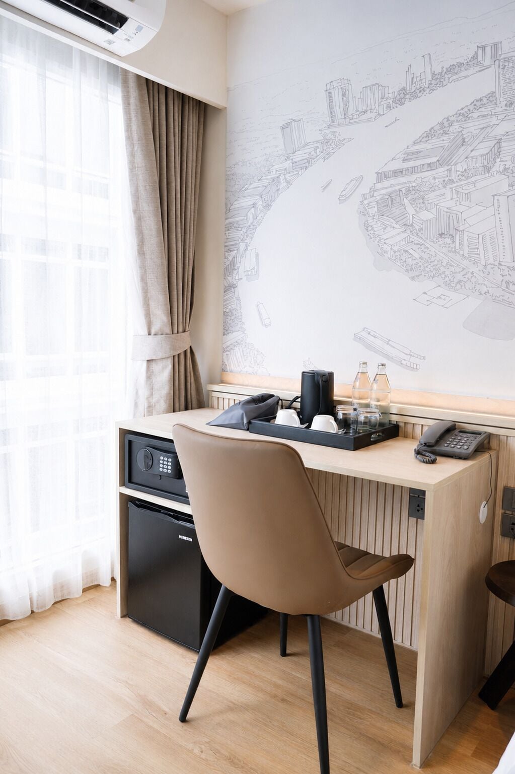 A hotel room with a desk, a chair, a coffee maker, a phone, and a wall with a cityscape mural.