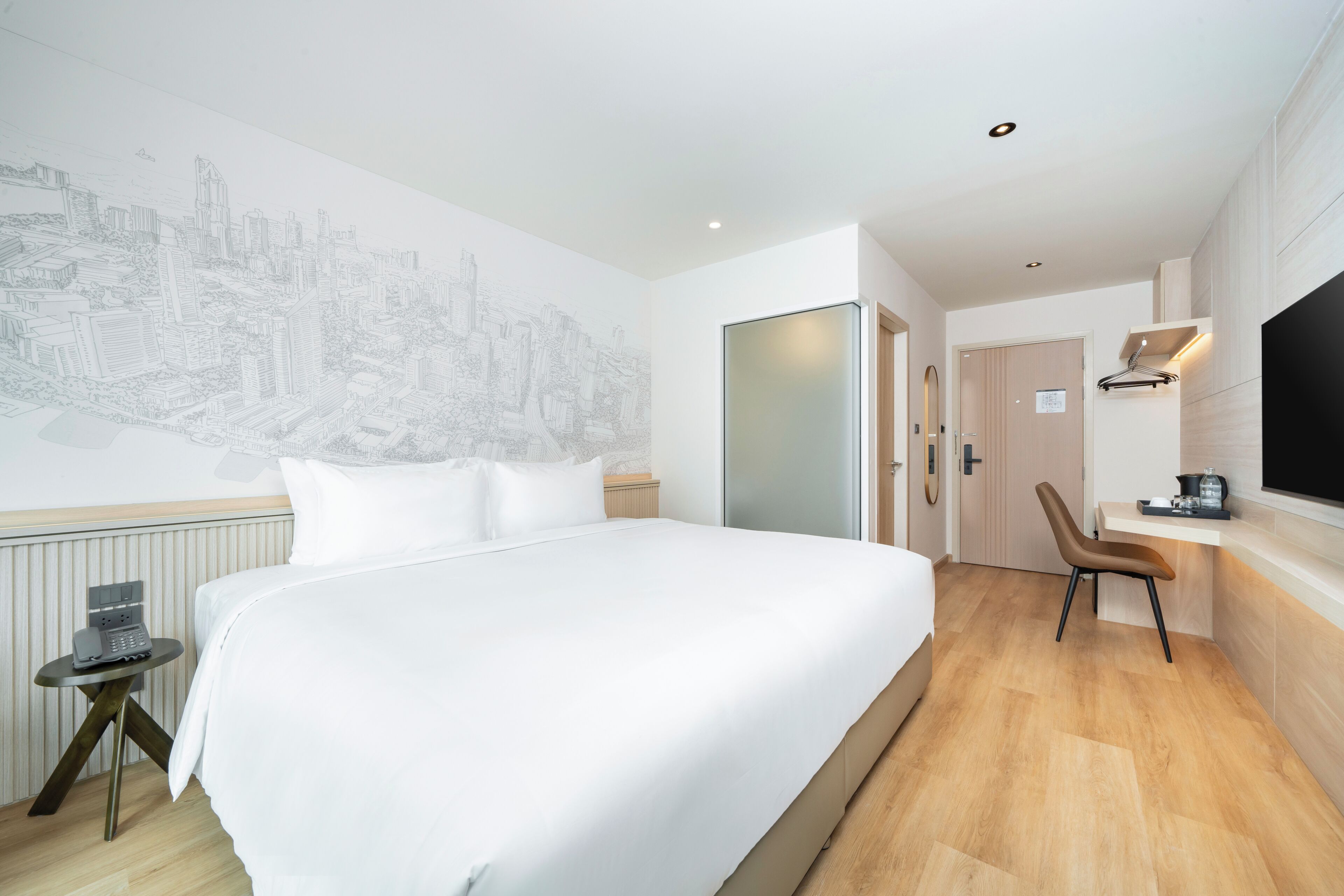 A hotel room with a large bed, a desk with a chair, a flat-screen TV, and a cityscape mural on the wall.