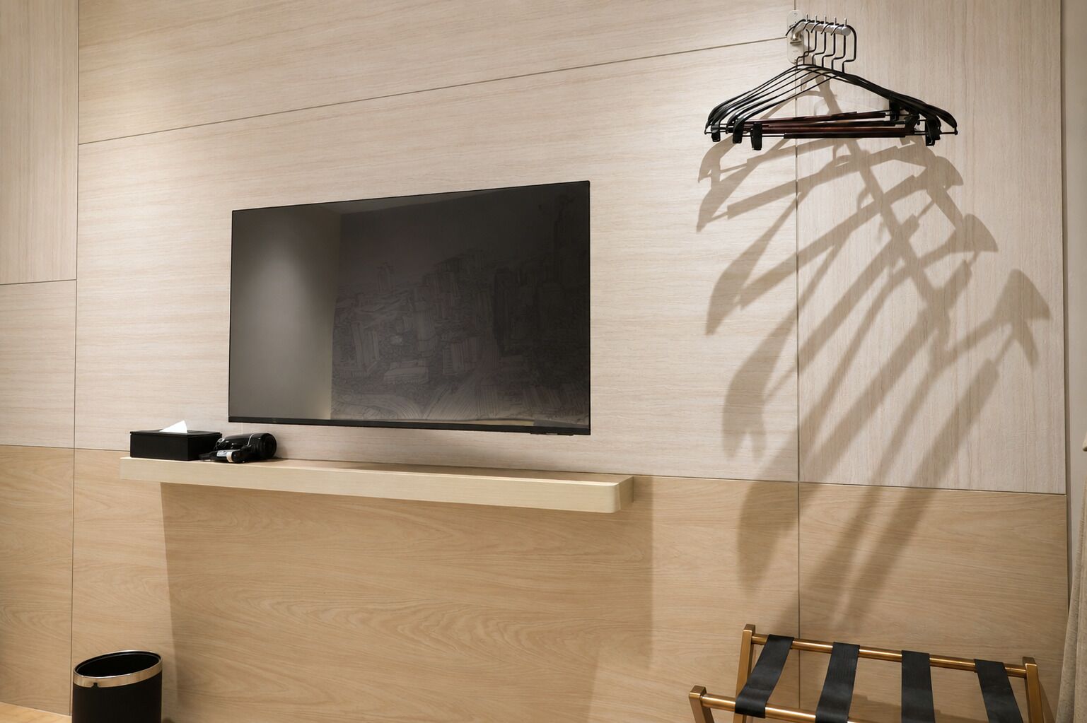 Quarto moderno com TV de tela plana presa a uma parede de madeira clara, uma prateleira com uma câmera, uma lixeira e um cabideiro.