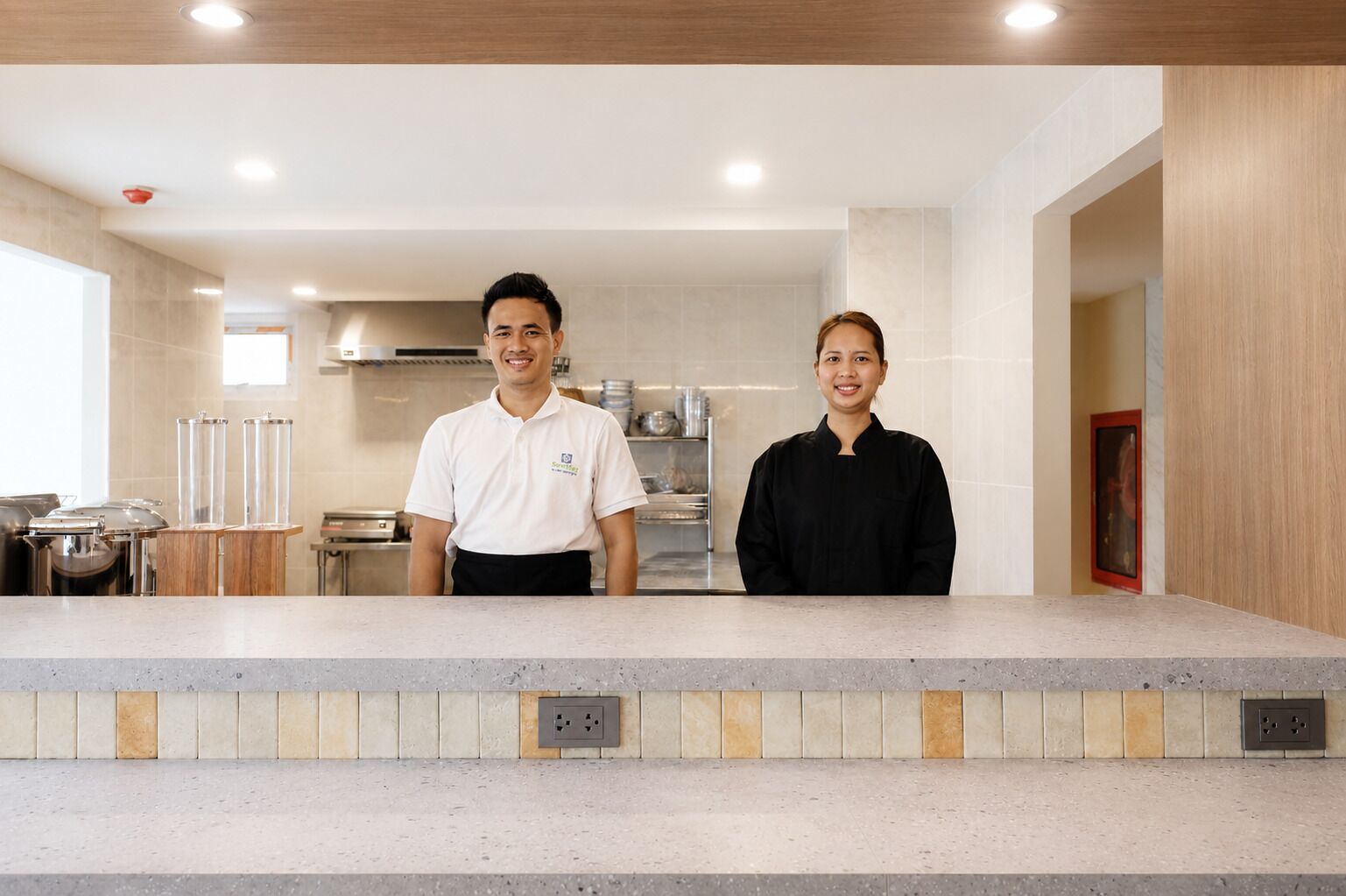 A modern kitchen with two individuals standing behind a counter, a tiled backsplash, and stainless steel appliances.