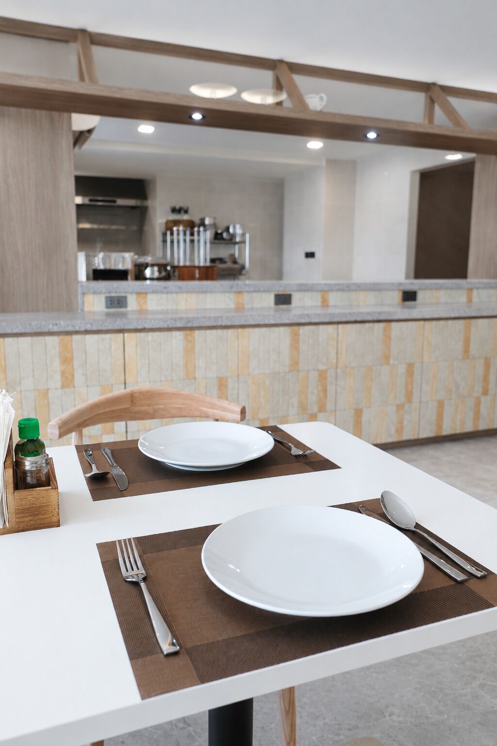 A dining table set with plates, cutlery, and a bottle, with a kitchen in the background.