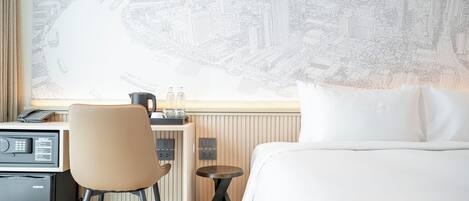 A hotel room with a bed, a desk with a chair, a small table, a kettle, and a wall art of a cityscape.