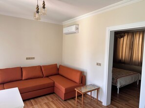 En stue med sofa, et lille bord, en aircondition og en seng i det tilstødende rum.