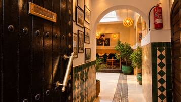 A hotel lobby with a dark wooden door, a fire extinguisher, framed pictures on the wall, and a tiled floor.