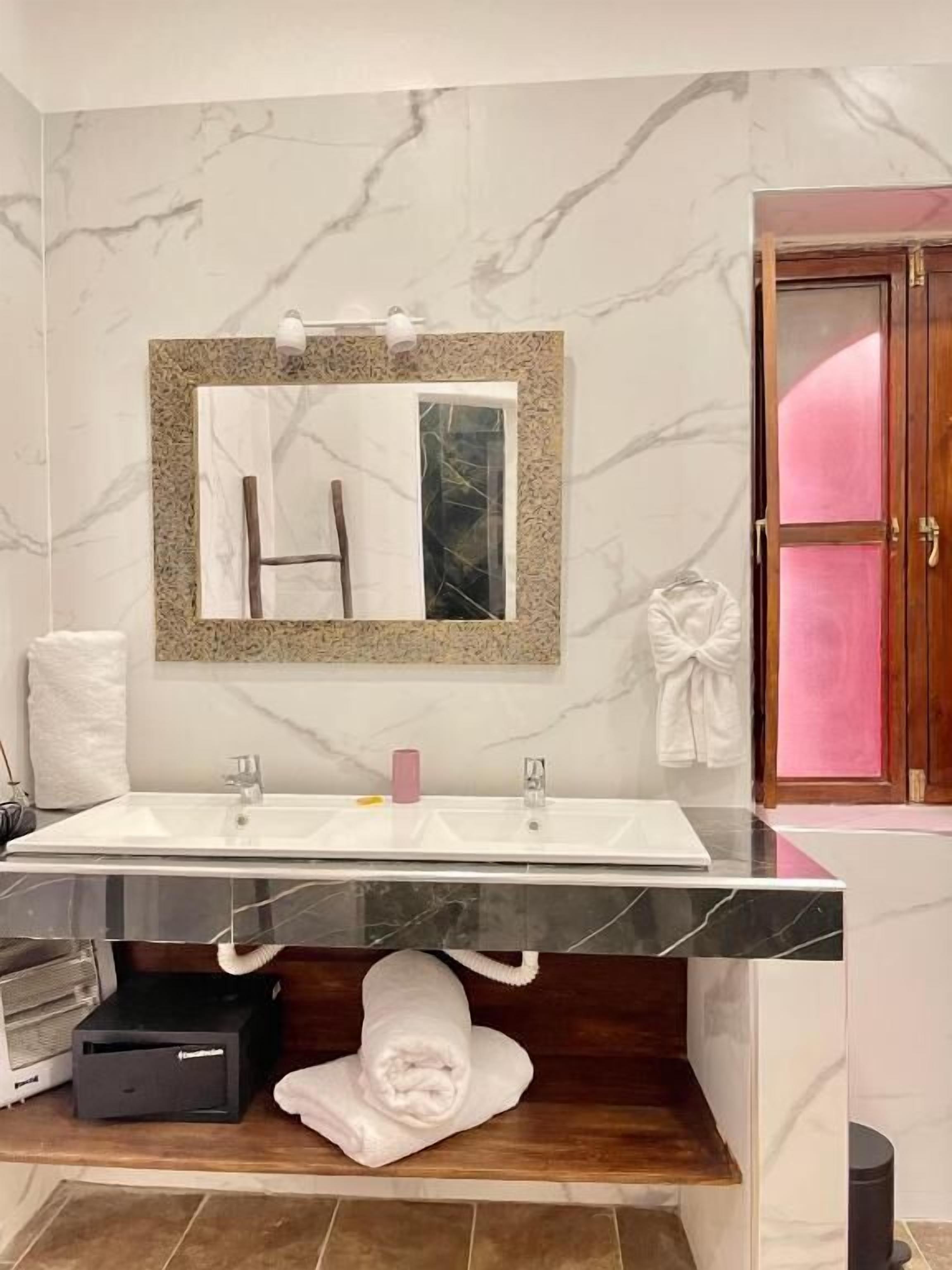 A bathroom with a marble wall, a sink with a mirror, a towel rack, and a wooden shelf.