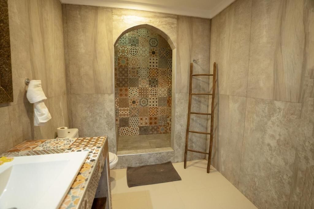 A bathroom with a tiled shower, a wooden ladder, a toilet, and a sink with a patterned tile backsplash.