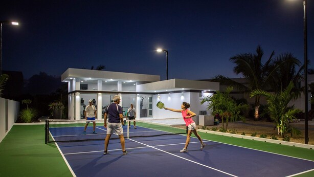 Ein Tennisplatz im Freien mit Netz, umgeben von einem modernen Gebäude mit Glasfenstern und einer flachen Dachkonstruktion.