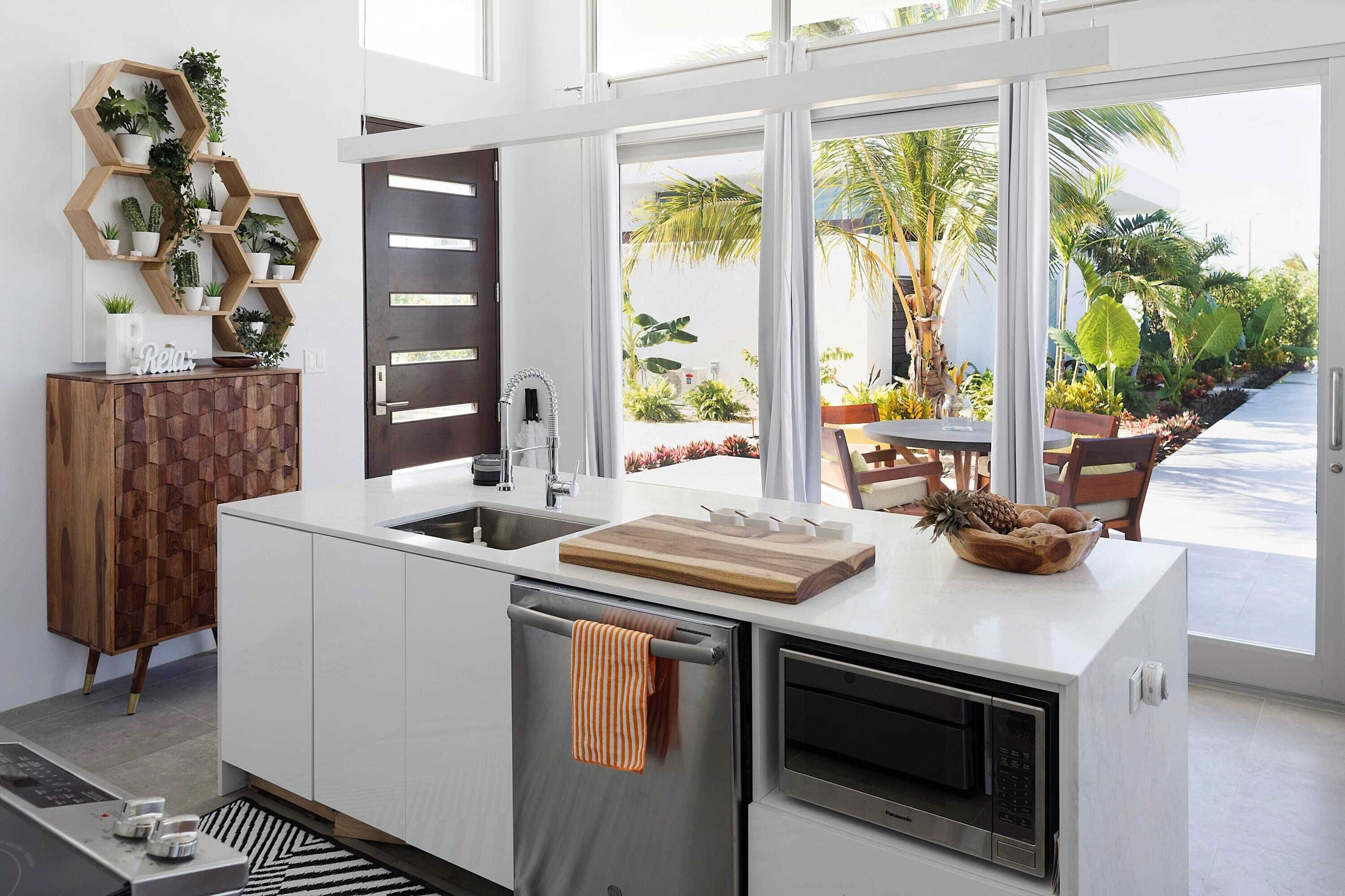 A modern kitchen with a built-in oven, a wooden cutting board, and a view of a tropical garden through large windows.