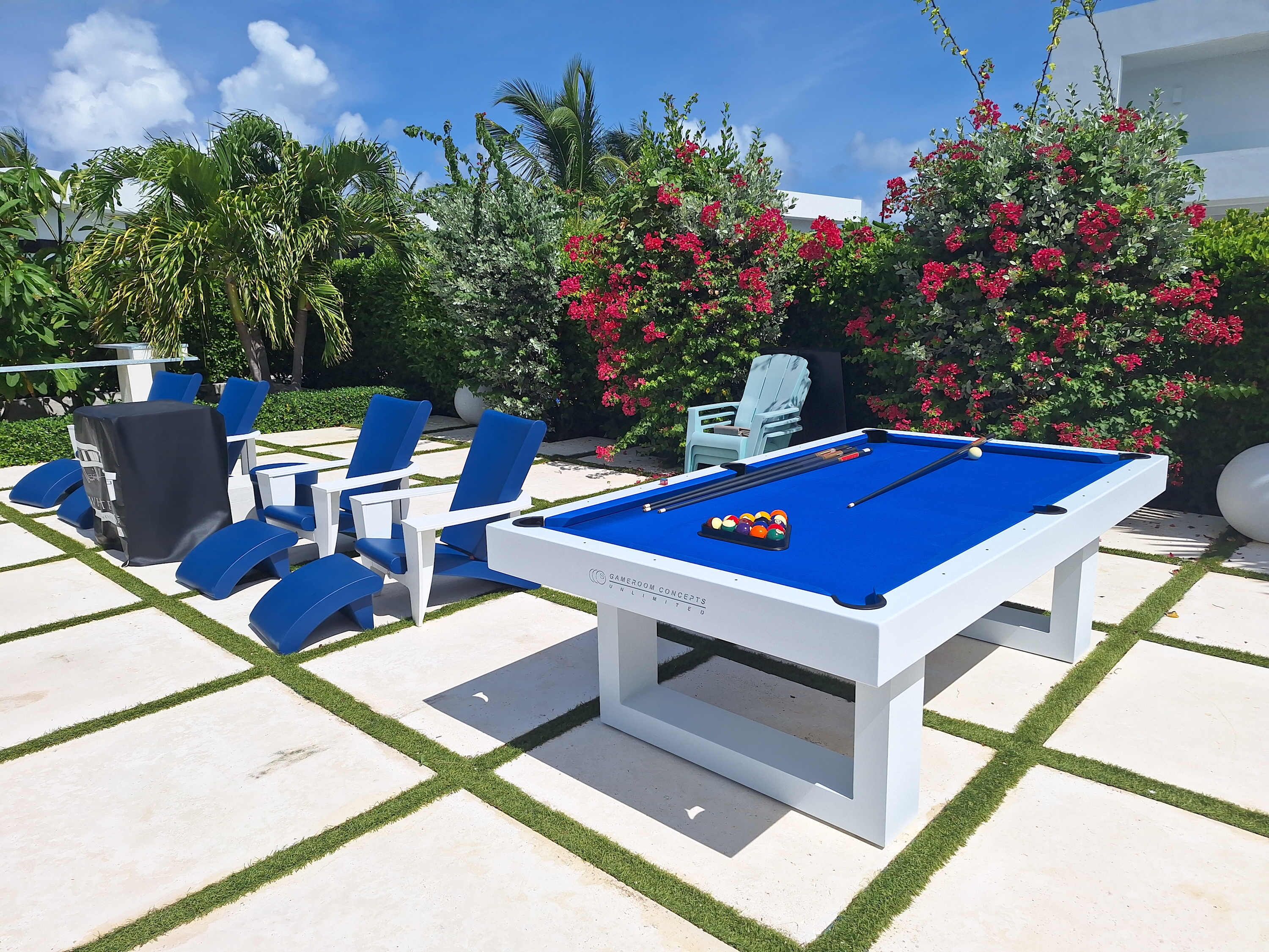 Een pooltafel met blauwe vilt, omringd door blauwe en witte loungestoelen, op een betegeld terras met kunstgras.