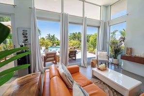 Una sala de estar moderna con un ventanal grande, un sofá, una mesa de centro y vistas a una piscina y palmeras.