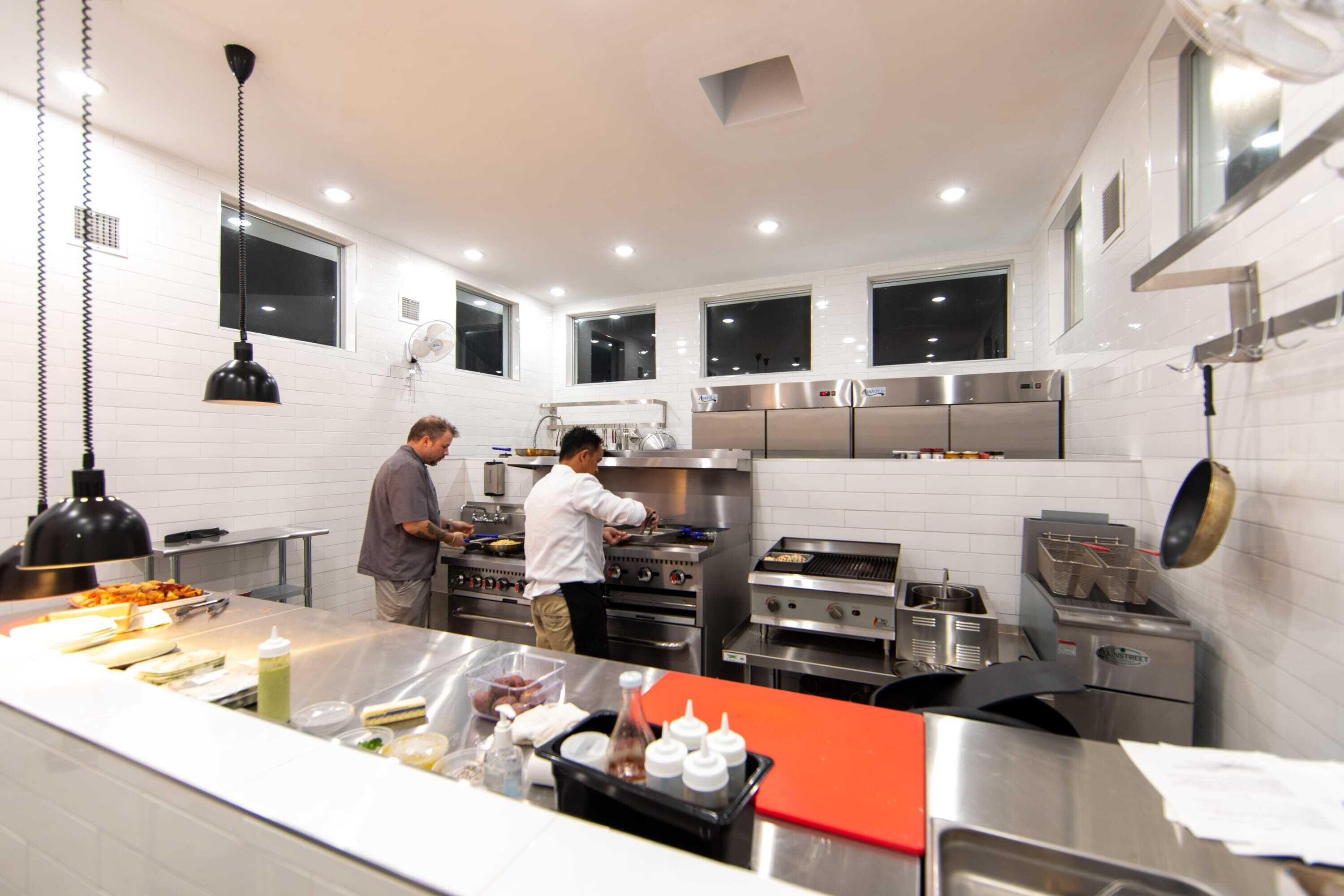 A modern kitchen with stainless steel appliances, a central island with food items, and two people cooking.