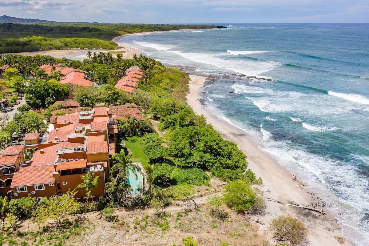 A coastal hotel with red-roofed buildings, a beach, and a pool.