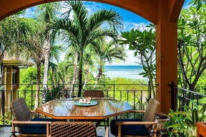 A balcony with a glass table, wicker chairs, and a view of the ocean. - Beachfront 2-BR Condo with Saltwater Pool & Sunset Views (Playa Langosta)