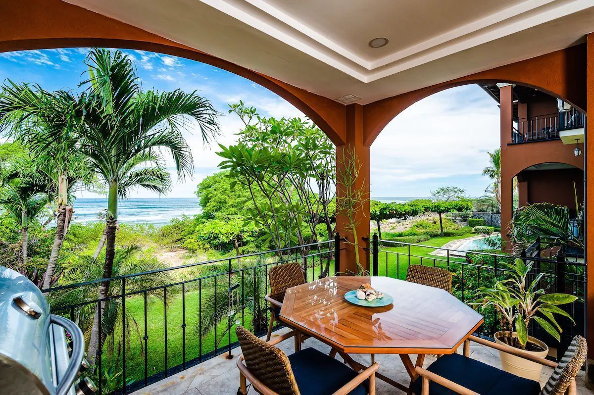 A balcony with a wooden table and chairs, overlooking a lush garden and the ocean.