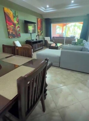 A living room with a dining table, sofas, a TV, and artwork on the walls. - Beachfront 2-BR Condo with Saltwater Pool & Sunset Views (Playa Langosta)