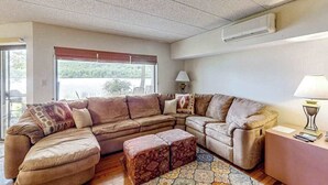 Ein Wohnzimmer mit einer Eckcouch, einem gemusterten Hocker, einer Lampe und Blick durch ein Fenster.