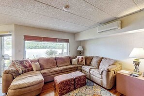 Ein Wohnzimmer mit einer Eckcouch, einem gemusterten Hocker, einer Lampe und Blick durch ein Fenster.