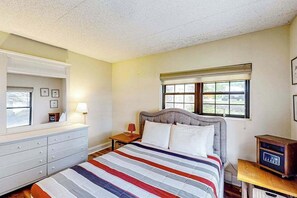 Ein Schlafzimmer mit Bett, Kommode, Nachttisch, Lampe, Fenster und einem Radio.