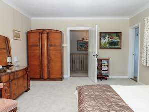 A bedroom with a large wooden wardrobe, a bed with a brown quilt, a dresser, a mirror, a painting, and a bookshelf.