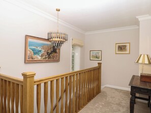 Un couloir avec une rambarde en bois, un lustre, des œuvres d’art encadrées et une lampe.