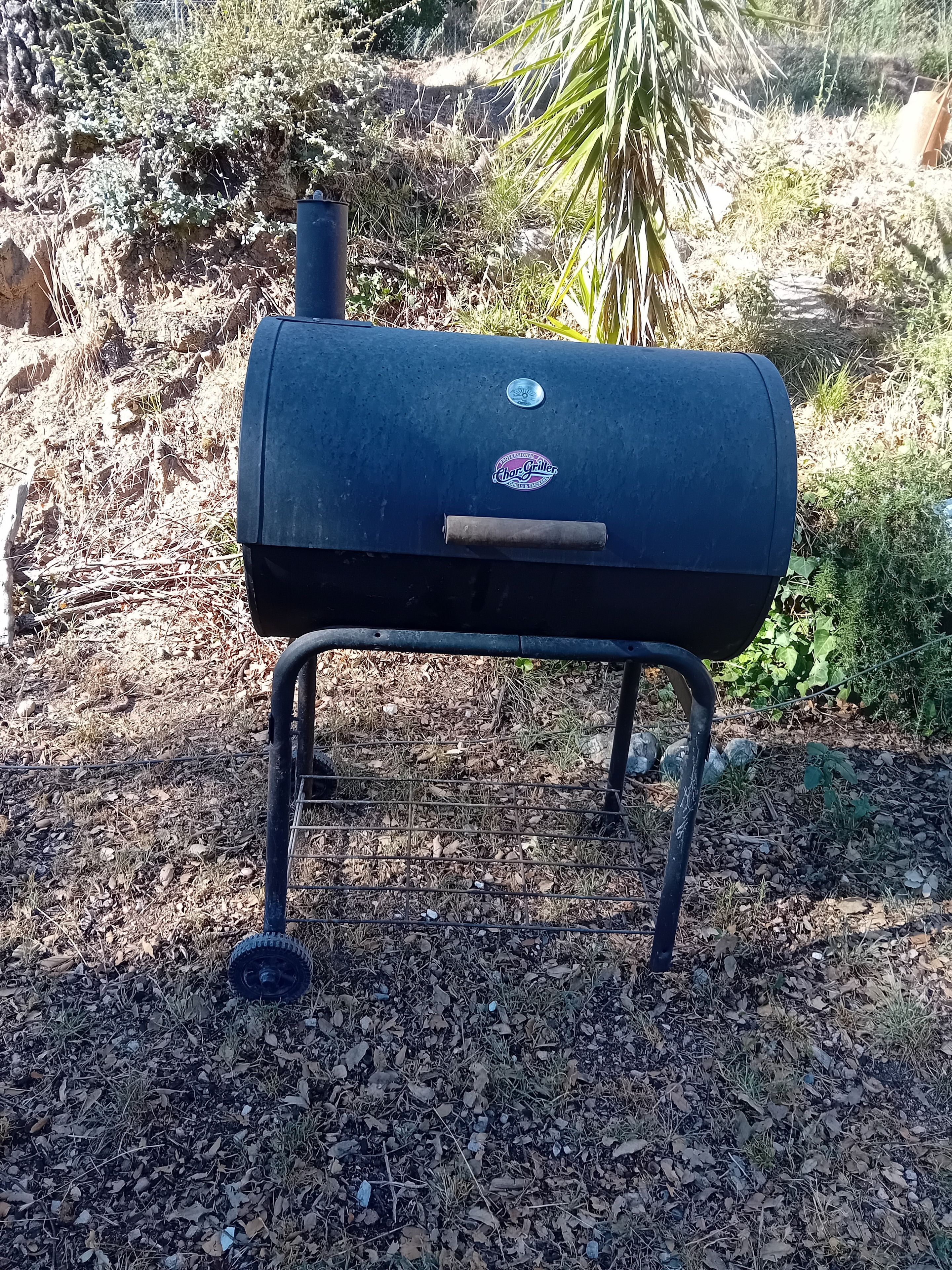 A charcoal grill with a logo, standing on a gravel surface with vegetation in the background.