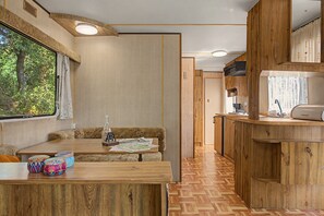 A compact RV interior with a kitchenette, a seating area with a table, and a window with a view of greenery.