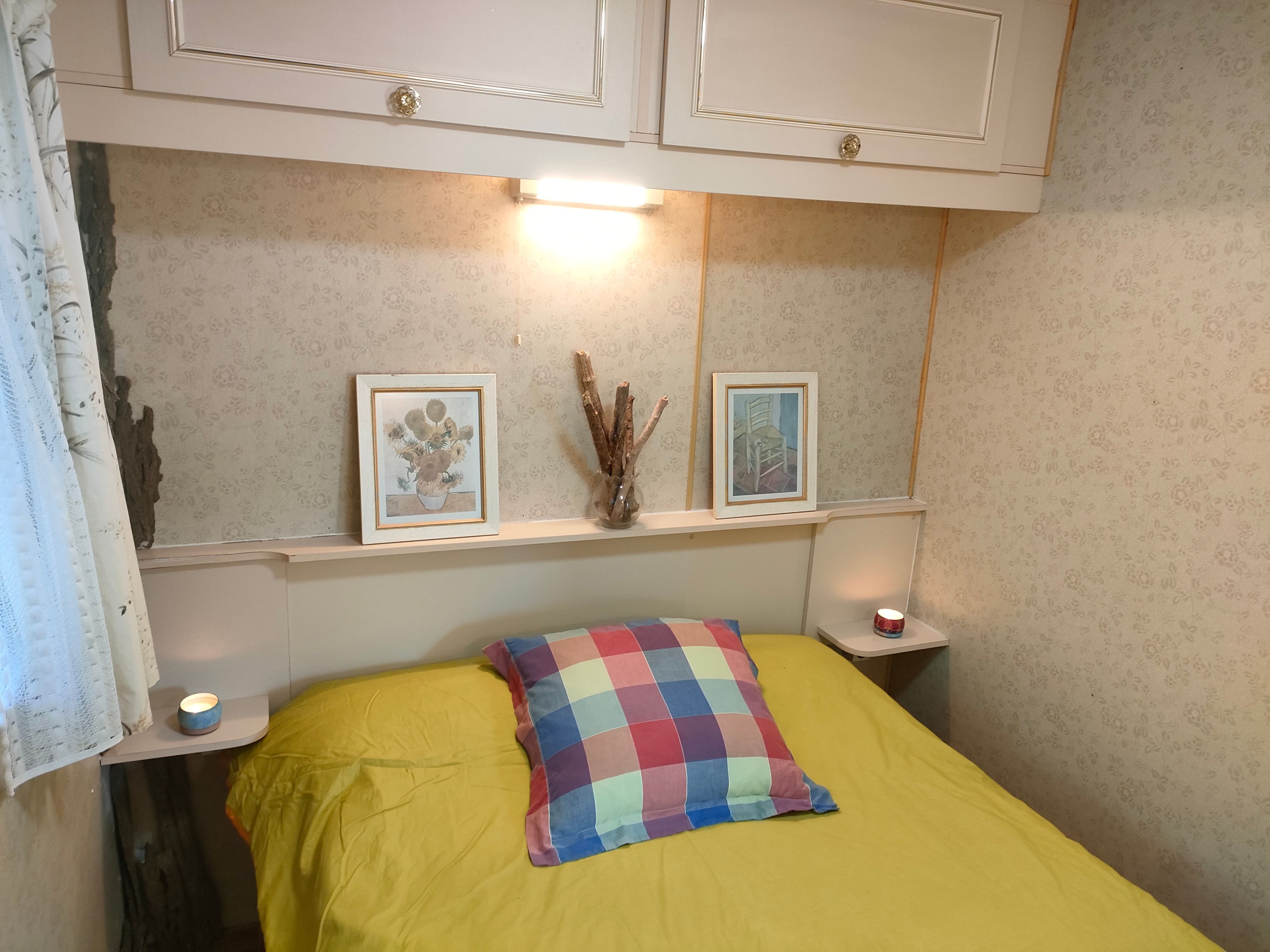 A small, cozy room with a bed, a nightstand, and a shelf with framed pictures.