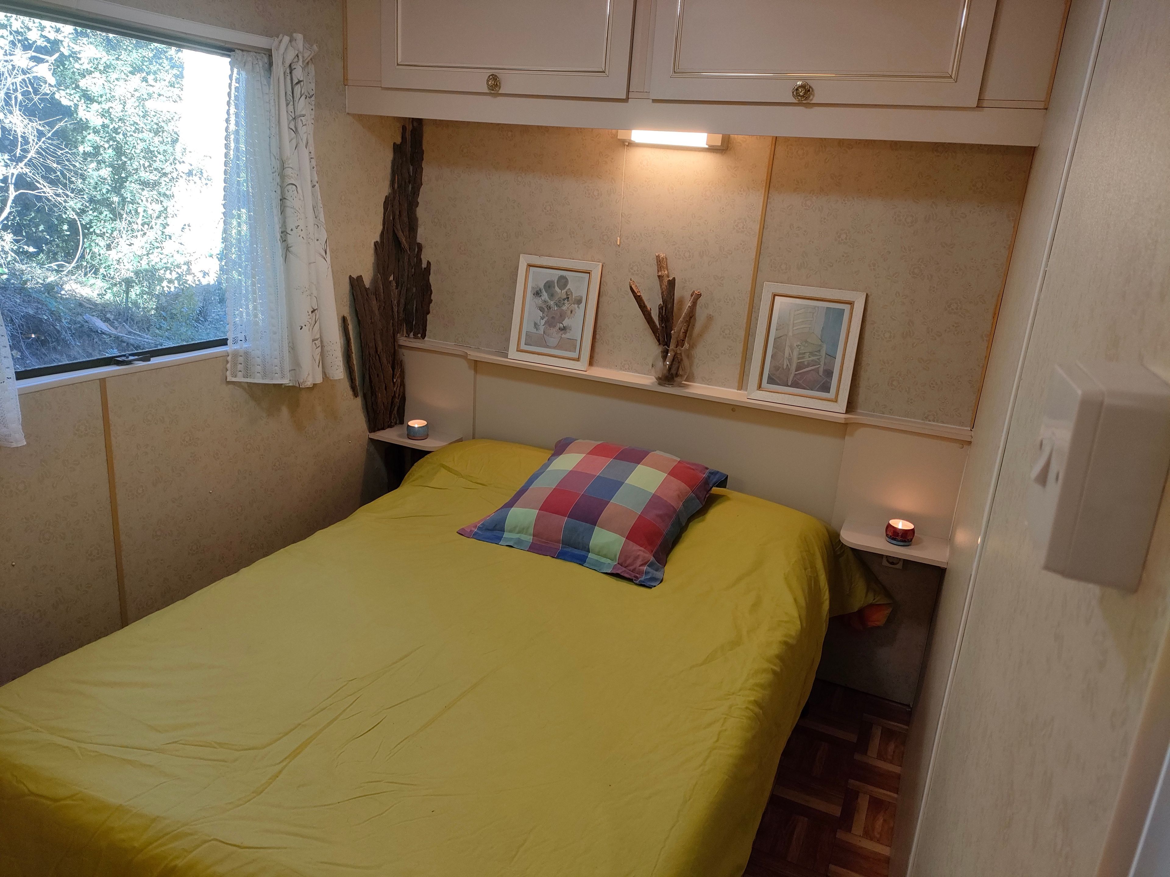 Un lit simple avec une couvre-lit jaune et un oreiller multicolore, flanqué de deux tables de chevet sur lesquelles sont posées des bougies, une fenêtre ornée de rideaux en dentelle et une étagère murale avec des photos encadrées et des branches décoratives.