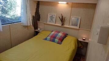 Un lit simple avec une couvre-lit jaune et un oreiller multicolore, flanqué de deux tables de chevet sur lesquelles sont posées des bougies, une fenêtre ornée de rideaux en dentelle et une étagère murale avec des photos encadrées et des branches décoratives.